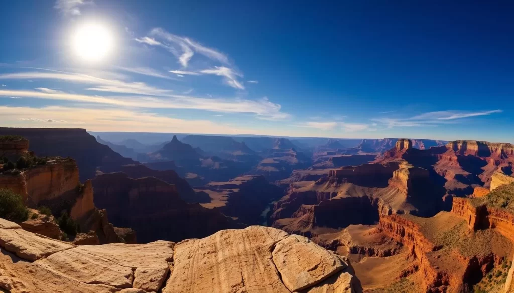 A breathtaking panoramic view of the iconic Grand Canyon South Rim, with its vast, layered multicolored rock formations stretching out to the horizon. The sun casts a warm, golden glow over the scene, highlighting the rugged, textured canyon walls and the winding Colorado River far below. In the foreground, a weather-worn rock outcrop provides a natural vantage point, inviting the viewer to take in the majestic, awe-inspiring landscape. Wispy clouds drift across the deep blue sky, adding depth and a sense of tranquility to the scene. The overall mood is one of serene, timeless grandeur, capturing the essence of this natural wonder.